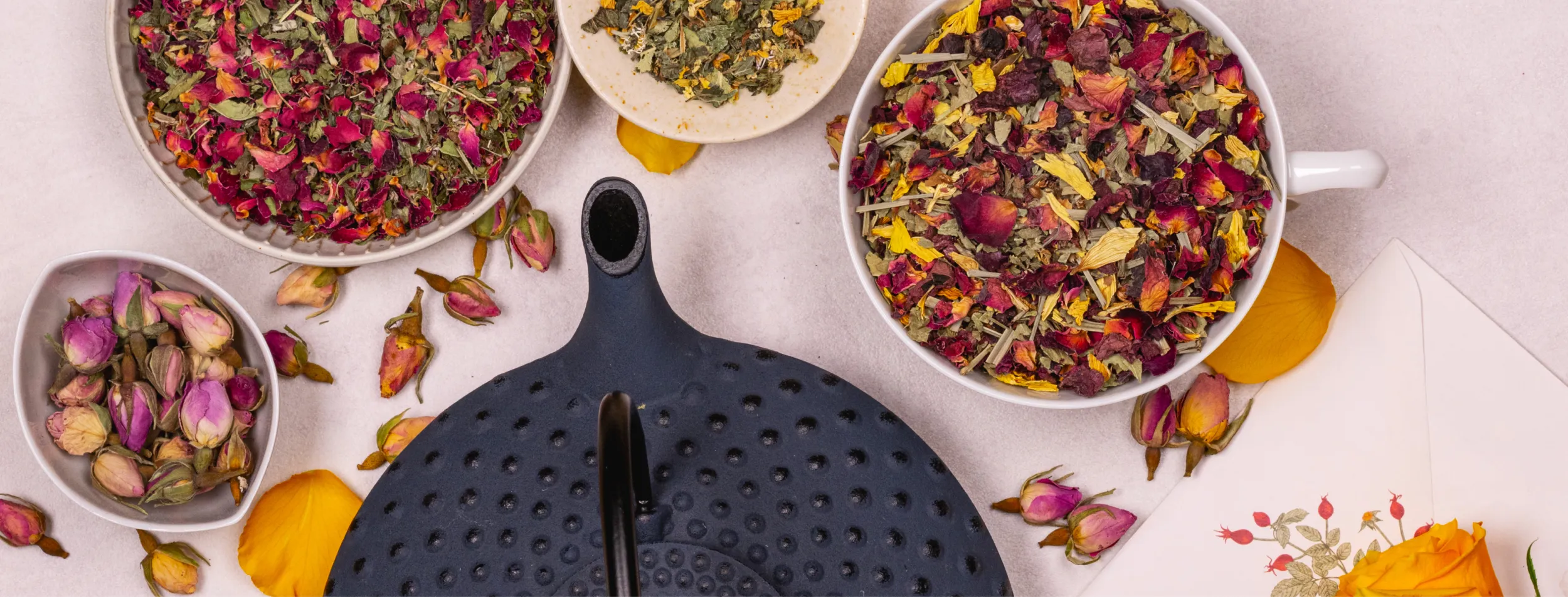 Dunkelblaue Teekanne aus Gusseisen umgeben von weißen Tassen und Schalen gefüllt mit Kräuter-Blütenteemischungen